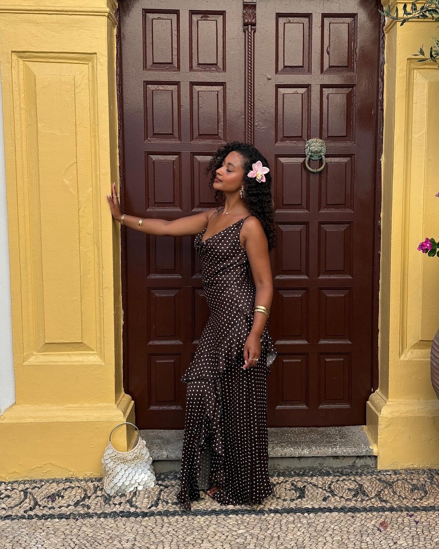 Fashion person Layla Abbey wears an Anthropologie dress in a recent image. She is infront of.a yelllow doorway with a brown wooden door. She is looking towards the door frame and has a pink lilly in her hair in a recent image from her Instagram.