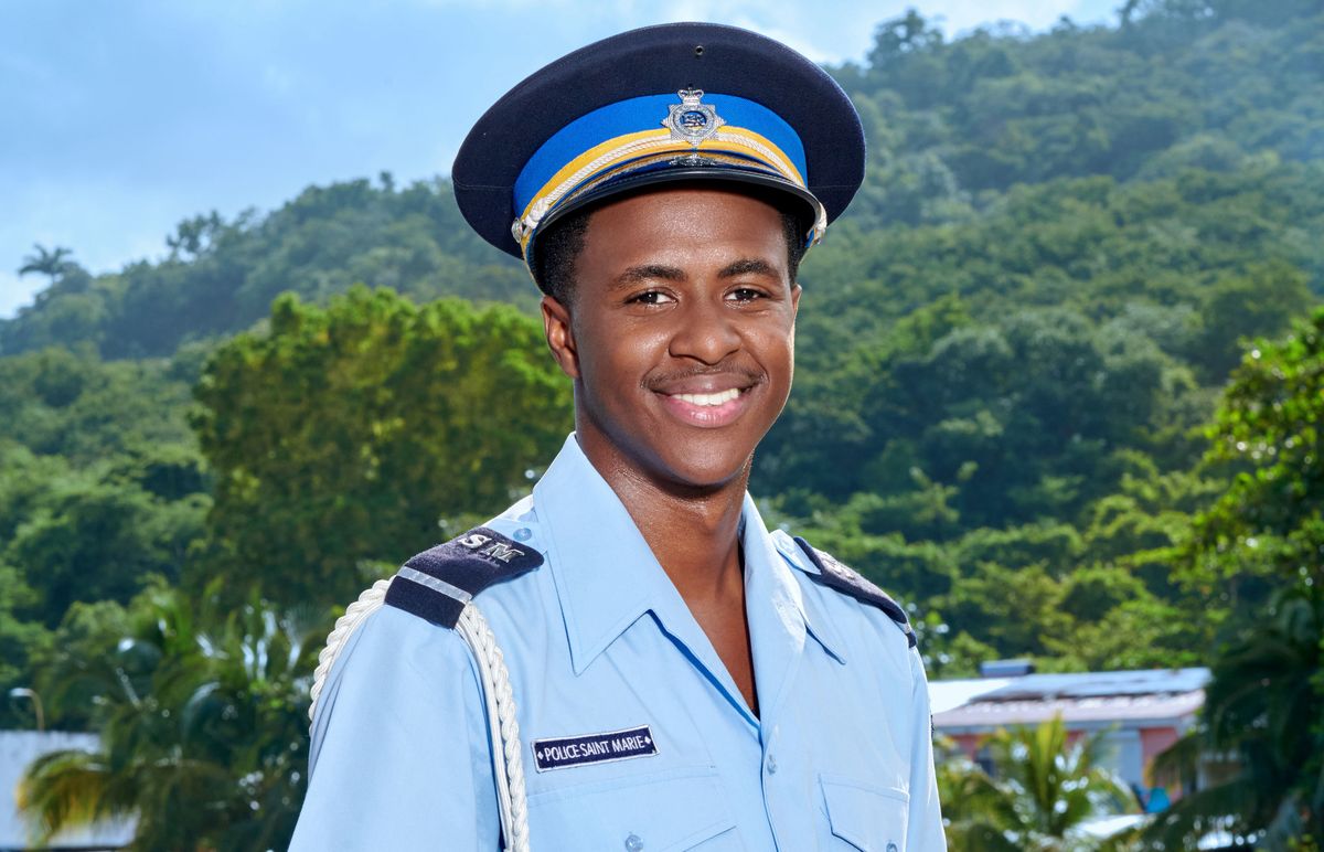 Death In Paradise&#039;s Officer Marlon Pryce (Tahj Miles) stands in his uniform in front of a bay and a verdant background of trees, smiling at the camera