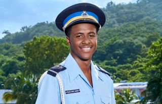 Death In Paradise&#039;s Officer Marlon Pryce (Tahj Miles) stands in his uniform in front of a bay and a verdant background of trees, smiling at the camera