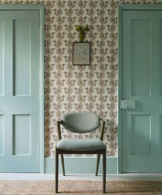 Hallway with floral wallpaper, blue painted doors and frames, chair placed in-between two doors, wooden frame, gray upholstery, white flooring, natural, textured rug