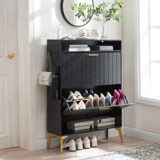 A black standing shoe cabinet. One drawer open showing pairs of sneakers and high heeled shoes. IN a white entryway. Bags hanging on the side.