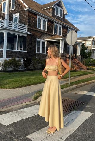 A woman wearing a yellow bandeau top with a matching maxi skirt, yellow pointed pumps, a raffia shoulder bag, and chunky gold bangles.