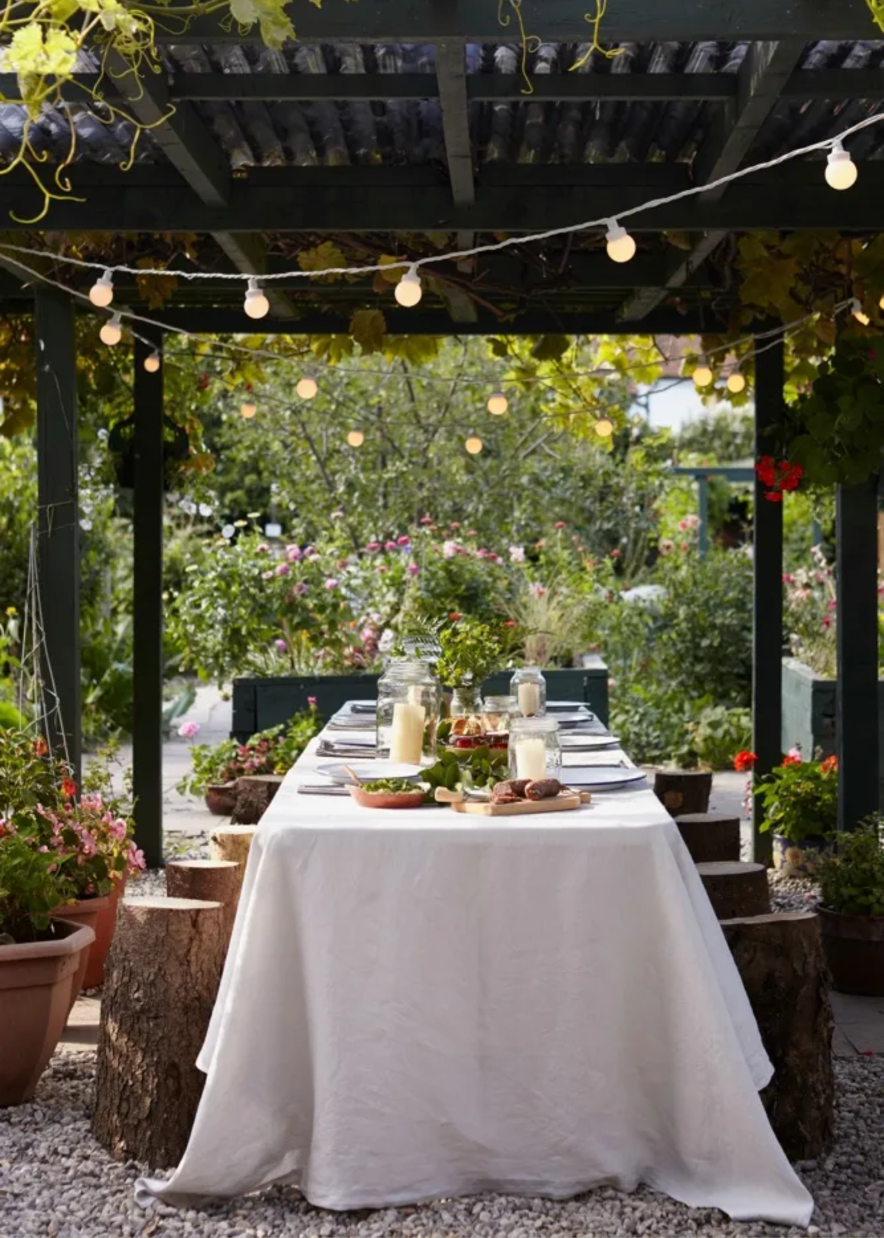 An outdoor dining table underneath a black pergola with tree stump stools, a white flowing tablecloth, and hanging string lights overhead
