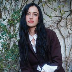 cazzie david poses against a wall with vines while wearing a black blazer and white shirt underneath