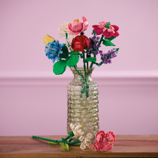 A bouquet of Aldi building block flowers on a wooden table in front of a pink background. There are roses, clover and lavendar in a clear ribbed vase.