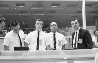 Standing at the flight director's console, viewing the Gemini 10 flight display in the Mission Control Center on July 18, 1966, are Glynn Lunney, prime flight director (second from left) with mission director William Schneider, Christopher Kraft, Manned Spacecraft Center director of flight operations; and Gemini program manager Charles Mathews.