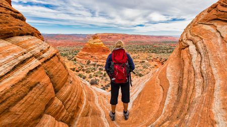 A woman wearing a red backpack stands on a colorful and swirly rock in Vermillion Cliffs National Monument in Arizona