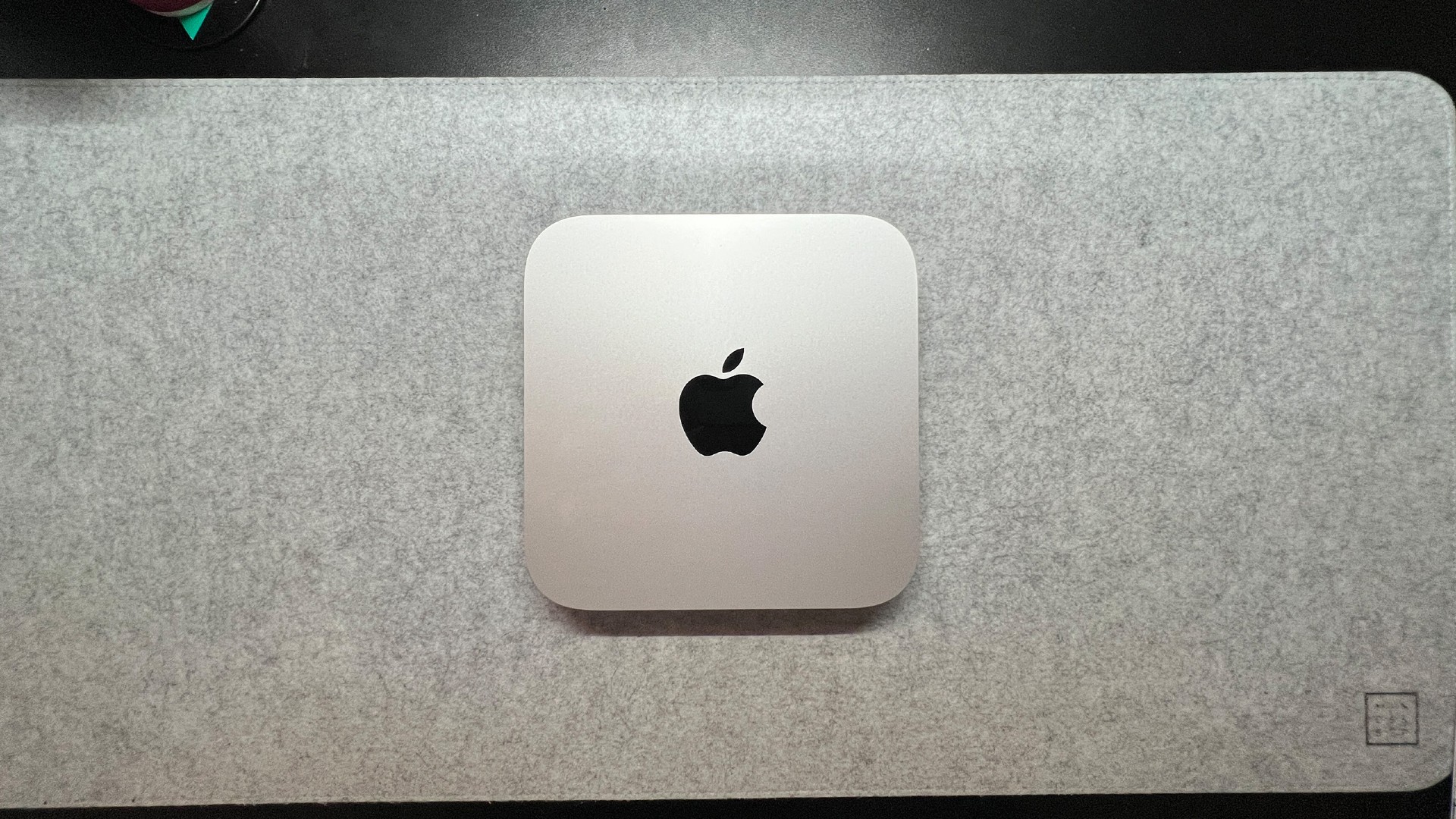 The Mac Mini Pro M2 is mini pc. It's a thin, square shape with rounded corners (20 x 20 x 3.5 cm). It's silver in color and has the Apple logo in the center of it (a black apple with a bite taken out of it). It is being displayed on a slightly darker gray computer desk mat.