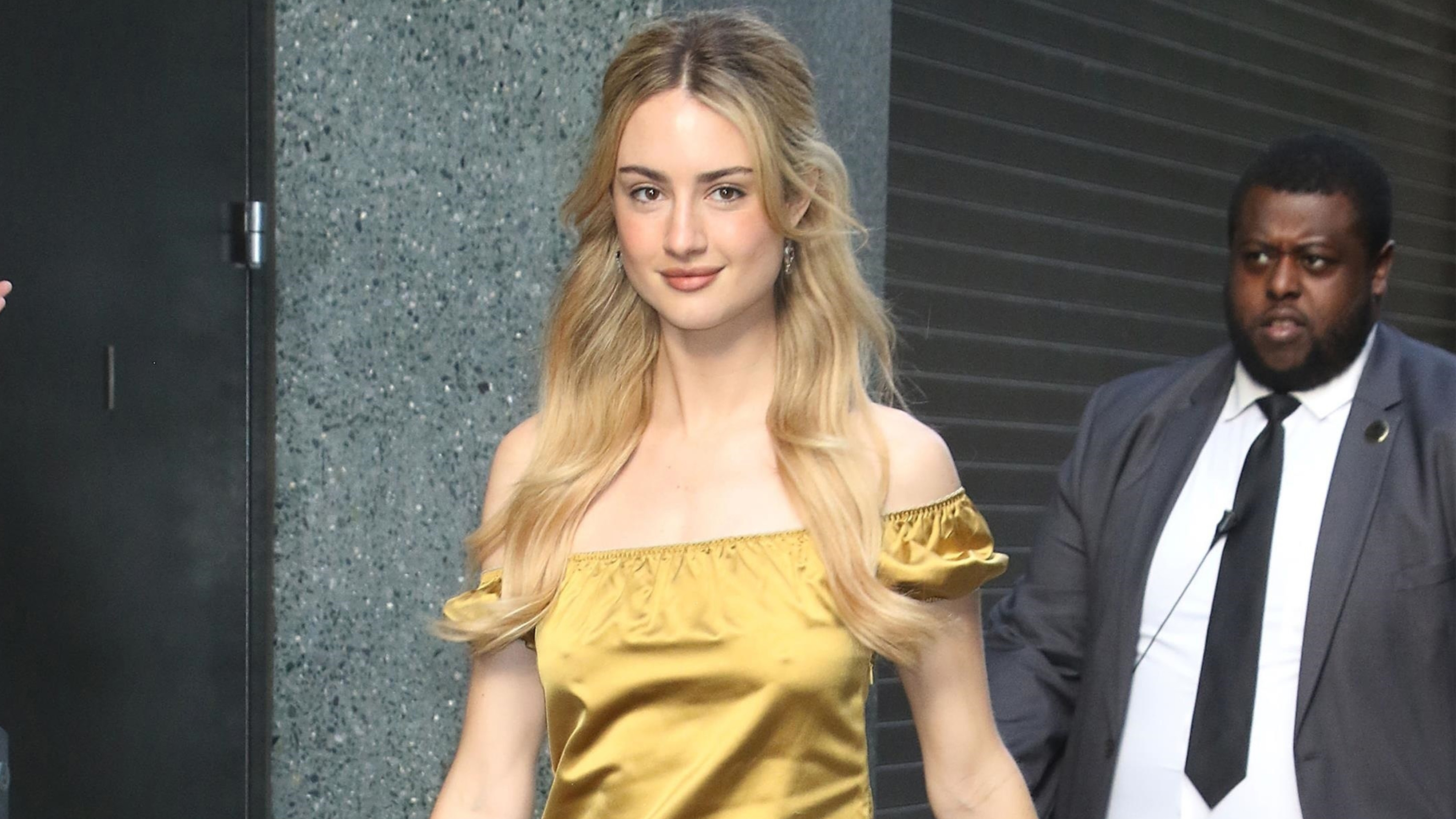 Grace Van Patten wears a gold satin minidress, black sheer tights, black heels, and a black leather bag.