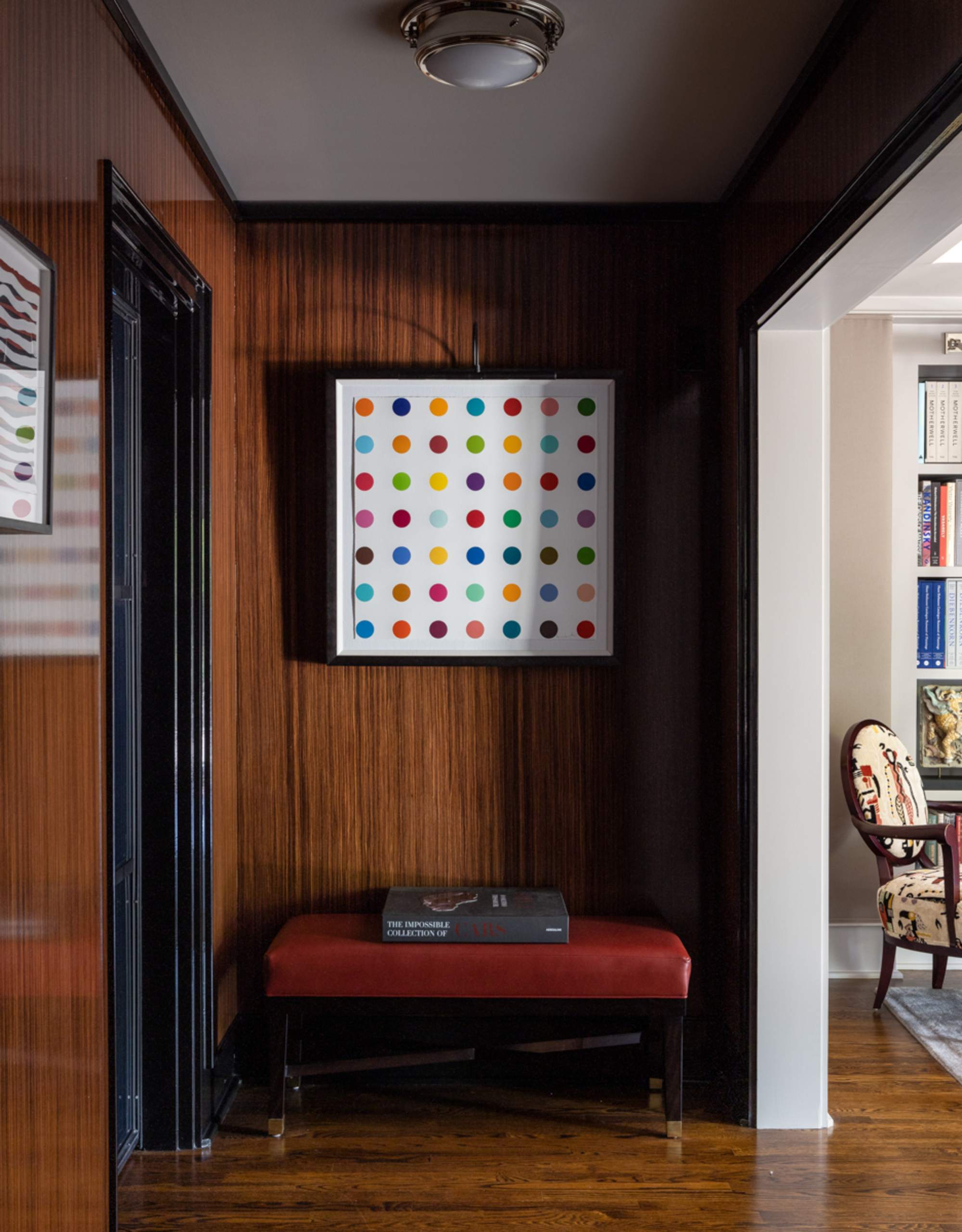 Image of a wood-drenched hallway with a red leather bench at the end and an artwork of multicolored polka dots on a white background hanging on the wall.