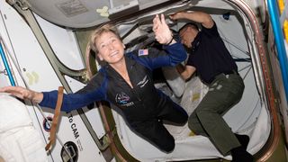 A woman in a black and blue flight suit floats through the hatch of a space station