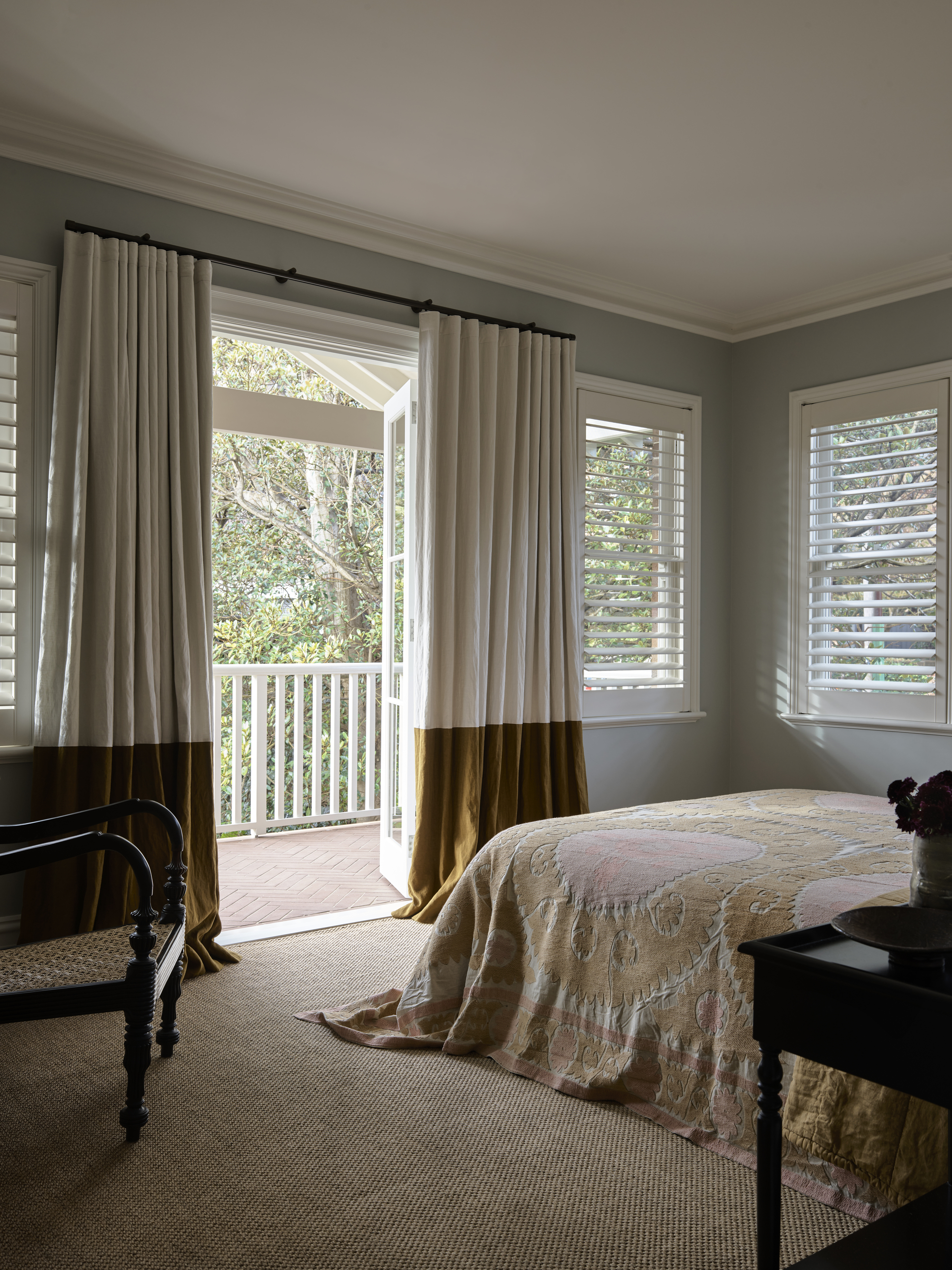 modern bedroom with sisal carpet, blue walls, white shutters on windows, white ceiling, antique chair in corner, bedcover, and two-tone curtains over doors to balcony
