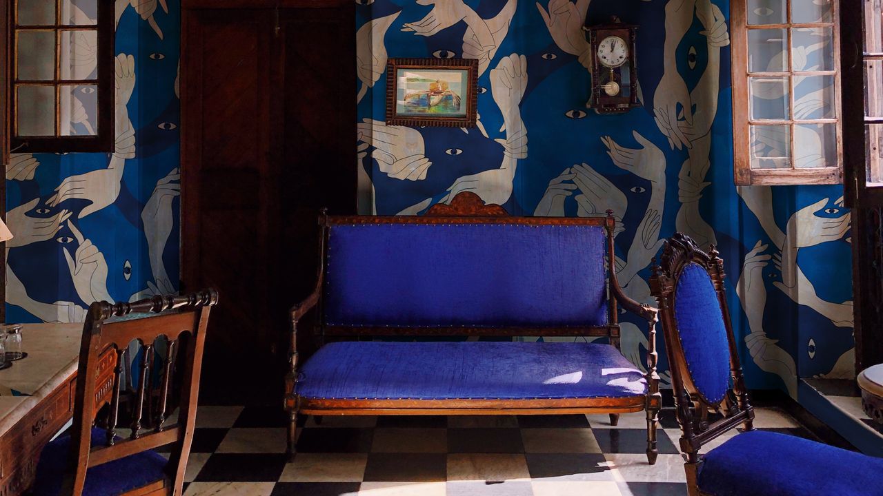 Image of a blue living room with an artistic blue and indigo wallpaper on the walls. There is a traditional indigo sofa with a wooden frame and a matching accent chair. The floors are made of a black-and-white checkered tile, and there is a wooden table and chair just in the frame.