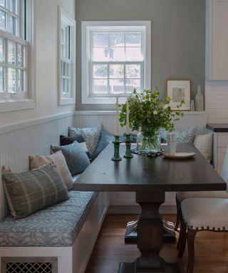 Corner dining room space with banquette bench style seating, two dining chairs, mahogany table, flowers in vase, candlestick, grey walls, blue and white seated cushions and chair pillows