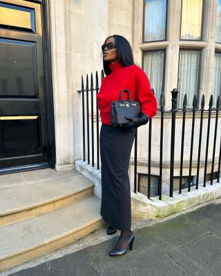 A woman wears a red sweater, black hermes bag, black pencil skirt, sheer tights, and black pumps.