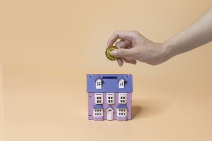 A small toy house with a large hand dropping coins into a slot in the roof.