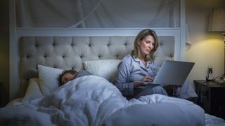 Lady works on her laptop while her husband sleeps next to her in bed