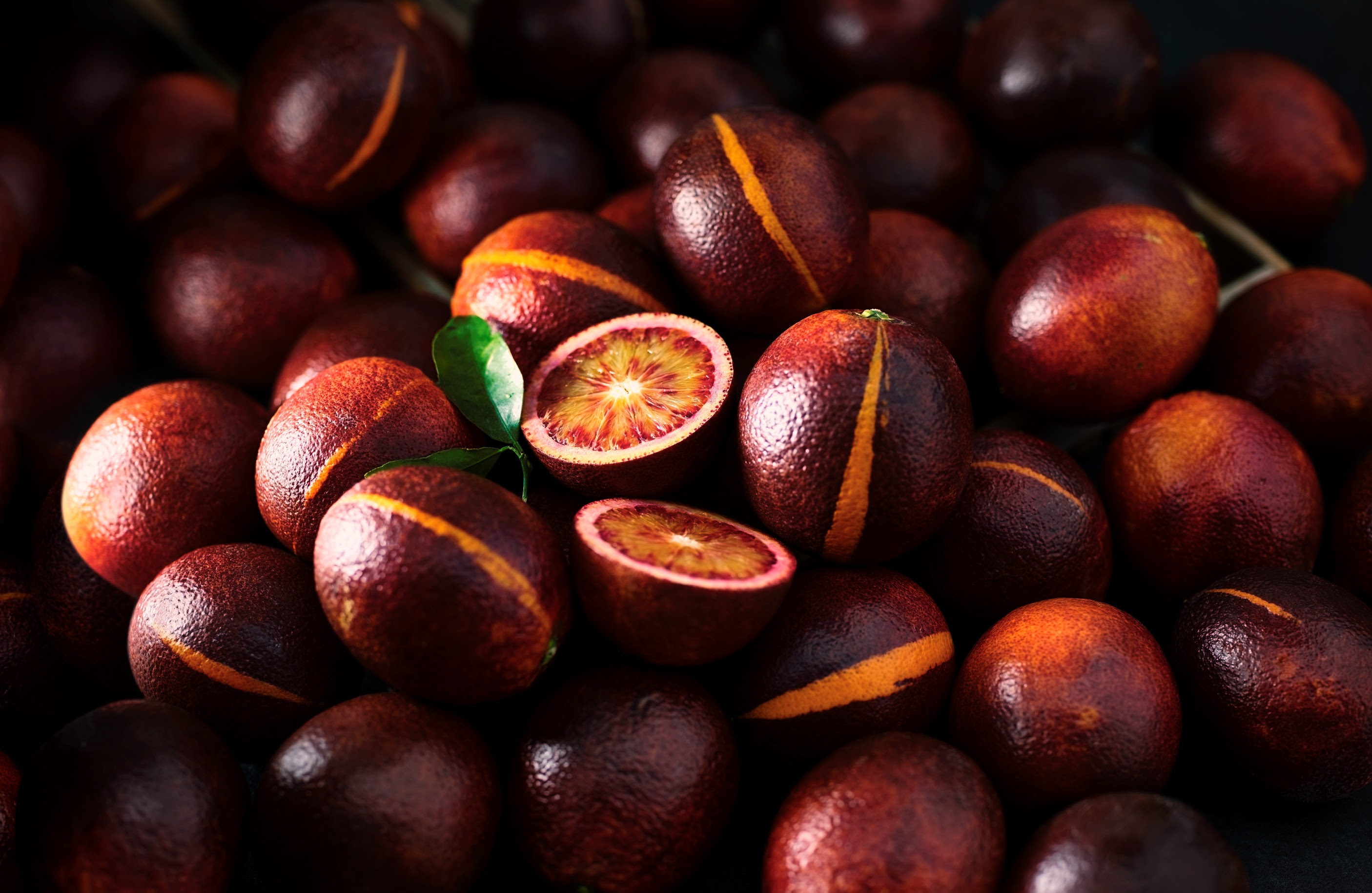 A dark, moody close-up of a pile of blood oranges, with a few cut open to reveal the striking red and orange interior.