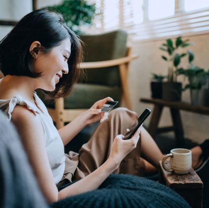 beautiful smiling young asian woman chilling at home, sitting on the floor in bedroom, enjoying a cup of coffee and shopping online with smartphone while making mobile payment with credit card on hand