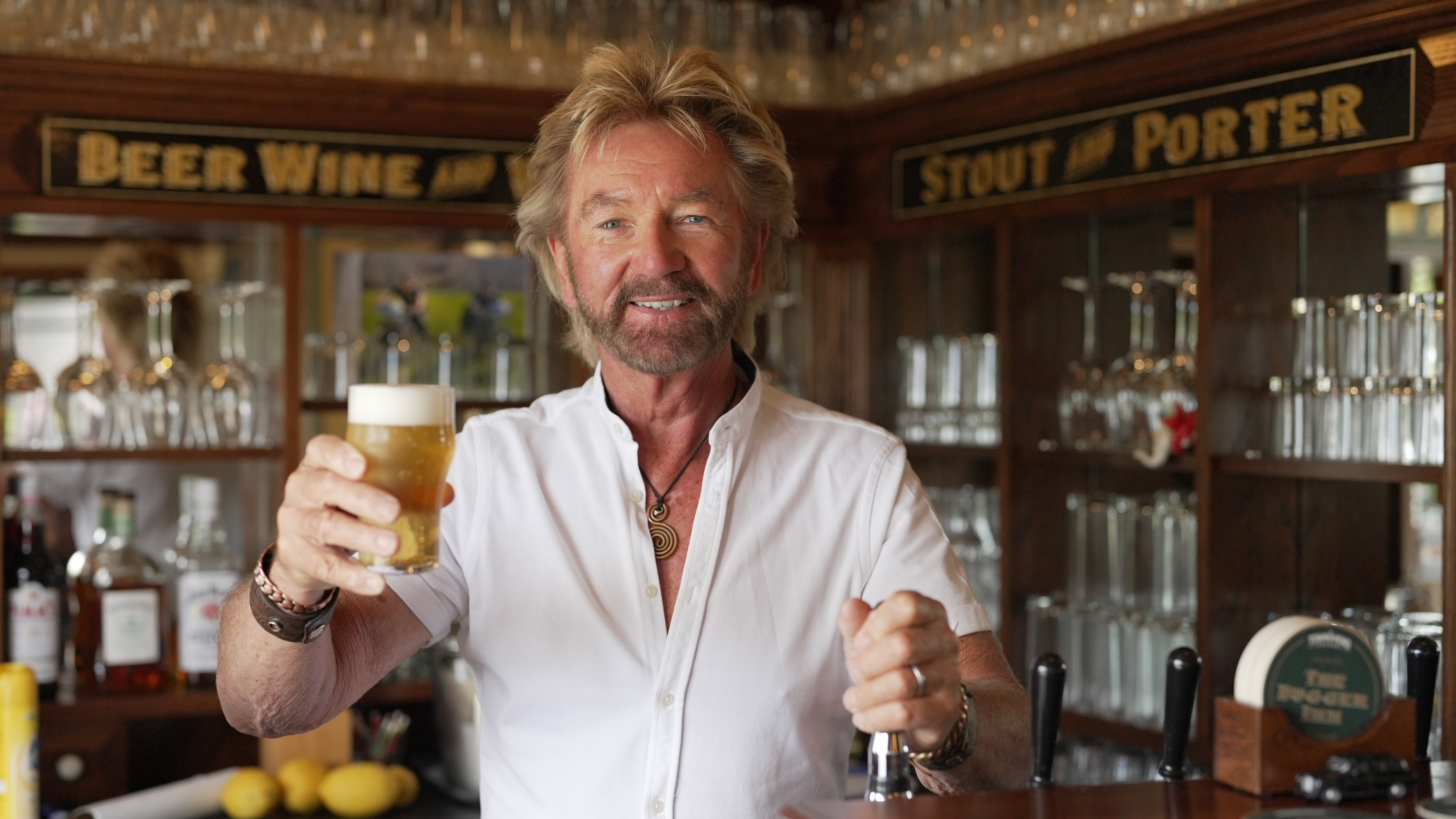 A smiling Noel Edmonds holds up a beer in his pub in New Zealand.