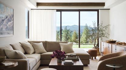 A living room space with large windows and sheer curtains. The living area has a large L shaped and an accent chair. There is also a beautiful view of the outdoor with large trees and green foliage 