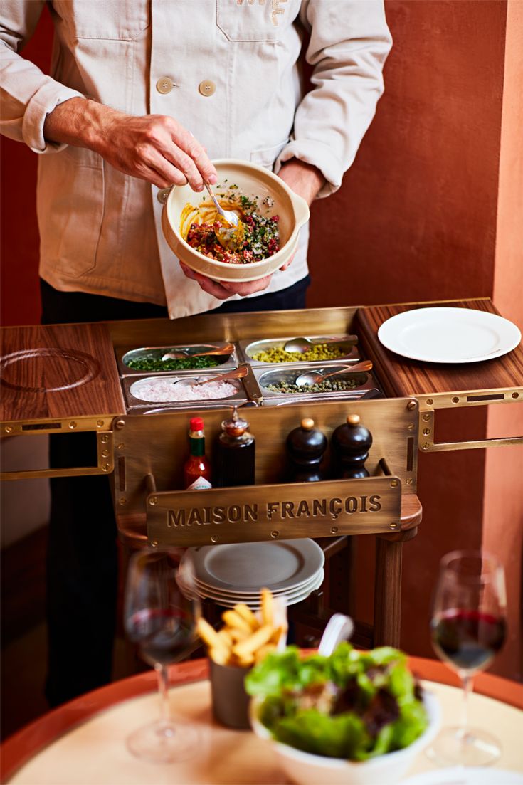 A waiter whizzing different ingredients tableside by picking toppings from a wood and brass branded kitchen trolley with the Maison Francois logo on it.