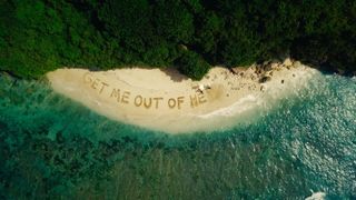 A birdseye view of a beach with the message 'Get Me Out of Here' written in the sand