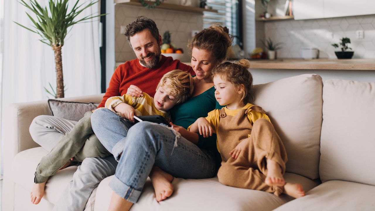 A young family of four look at a tablet while cuddled together on the sofa.