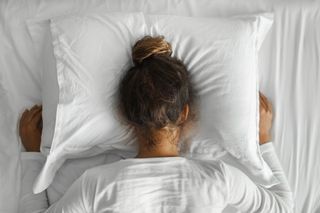 A woman with brown hair in a bun in bed with her head buried in the pillow