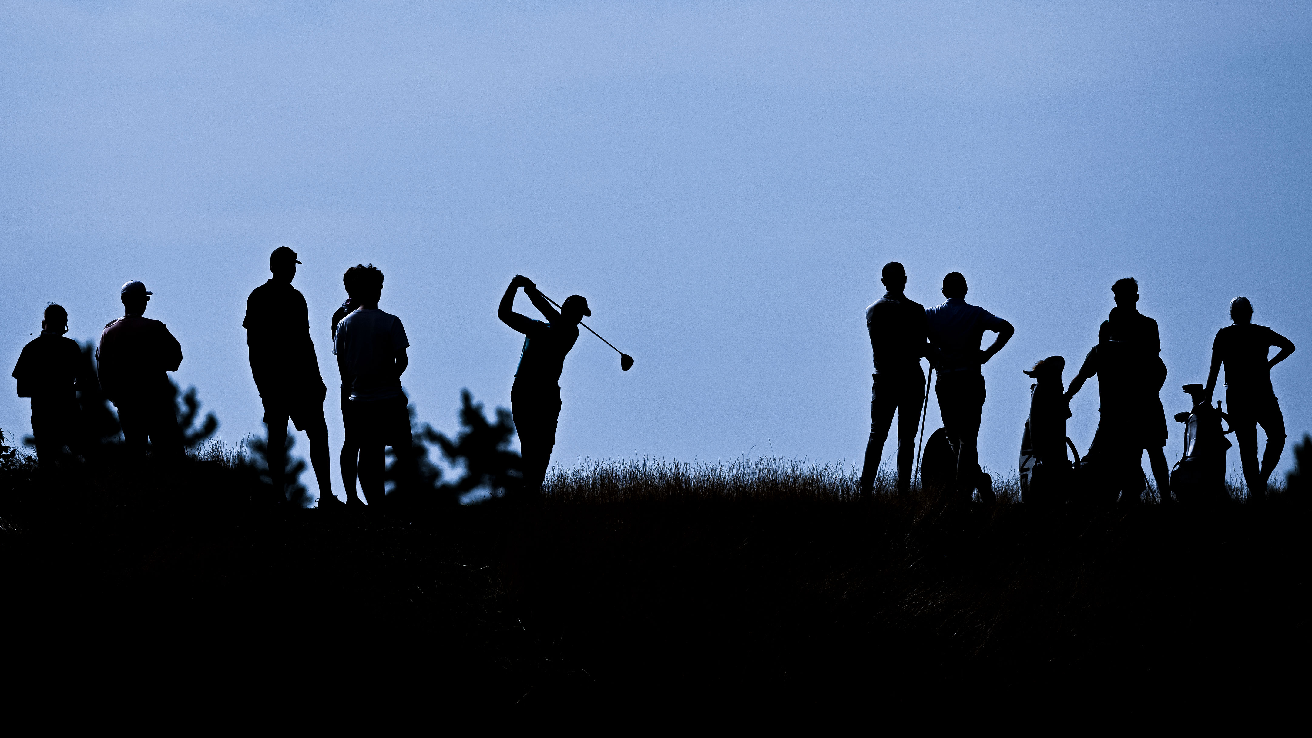 A group of silhouetted golfers watching one tee off 