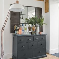 Dark grey chest of drawers with bar setup underneath mirror with industrial shutters beside oversized lamp