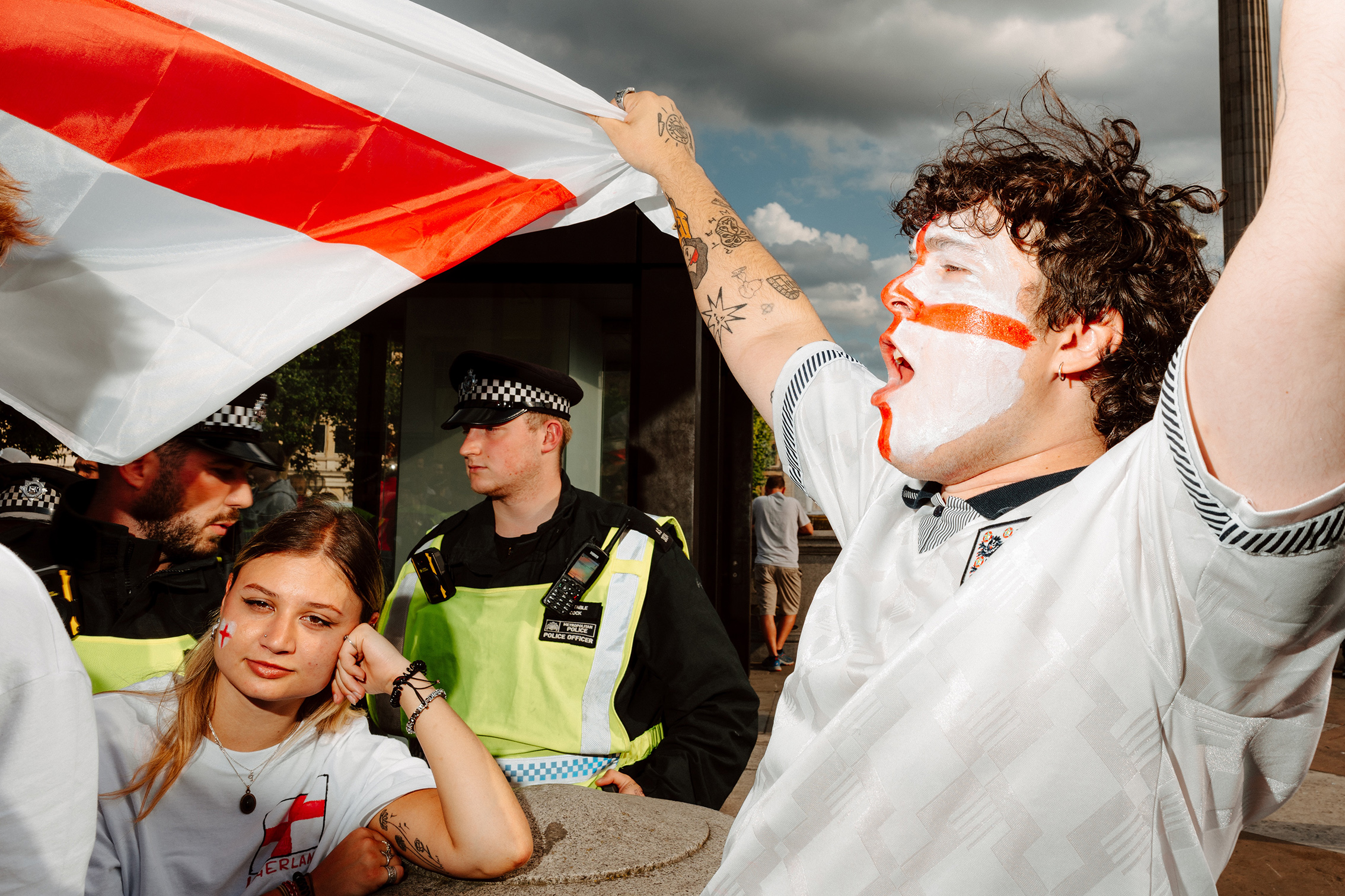 A man with face paint and tattoos cheers while holding an English flag. A woman looks thoughtful beside him. Two police officers stand in the background
