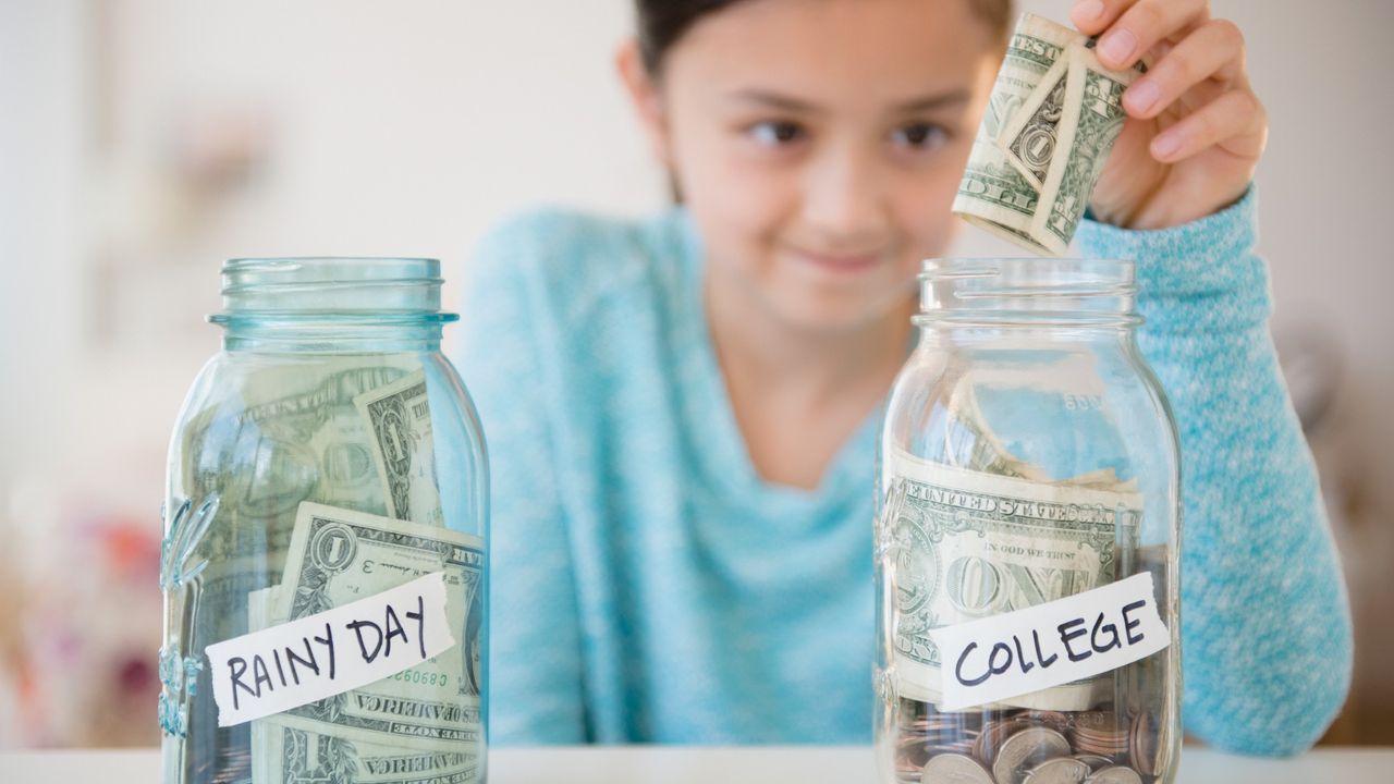 Little girl putting money into two jars, one that says &quot;Rainy day&quot; and one that says &quot;College&quot;