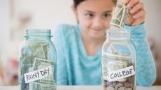 Little girl putting money into two jars, one that says "Rainy day" and one that says "College"
