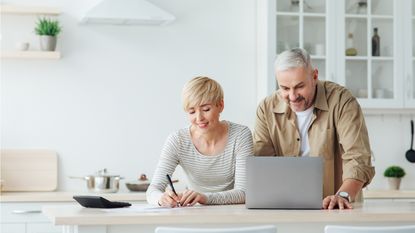 An older couple smile as they look over their investments together on a laptop on a kitchen island.