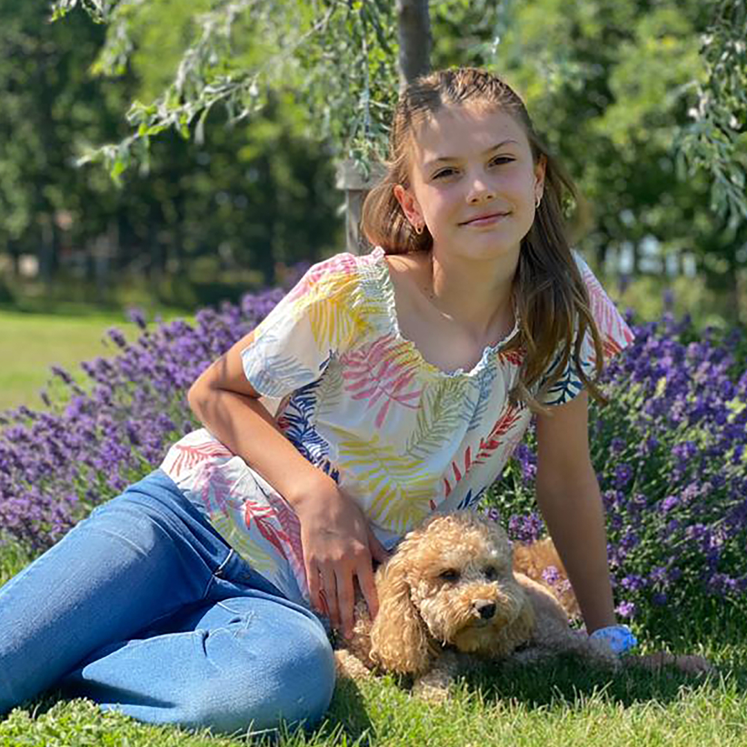 Princess Estelle wearing jeans and a colorful white shirt sitting in grass in front of purple flowers with a curly brown dog 