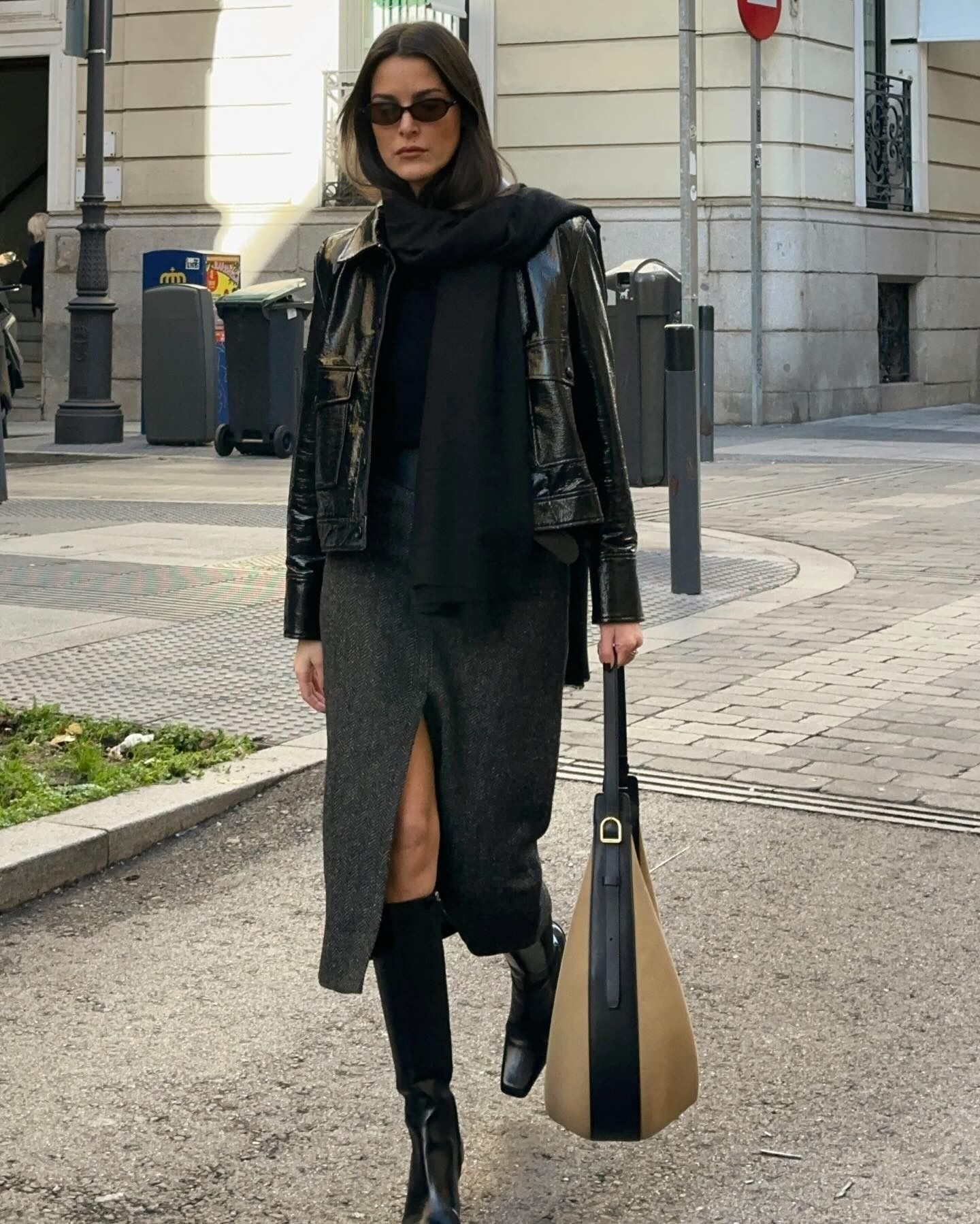 Influencer @leiasfez wears a patent leather jacket with a wool midi pencil skirt and black knee-high leather boots. She accessorises with sunglasses, a dark scarf and a light beige bag.