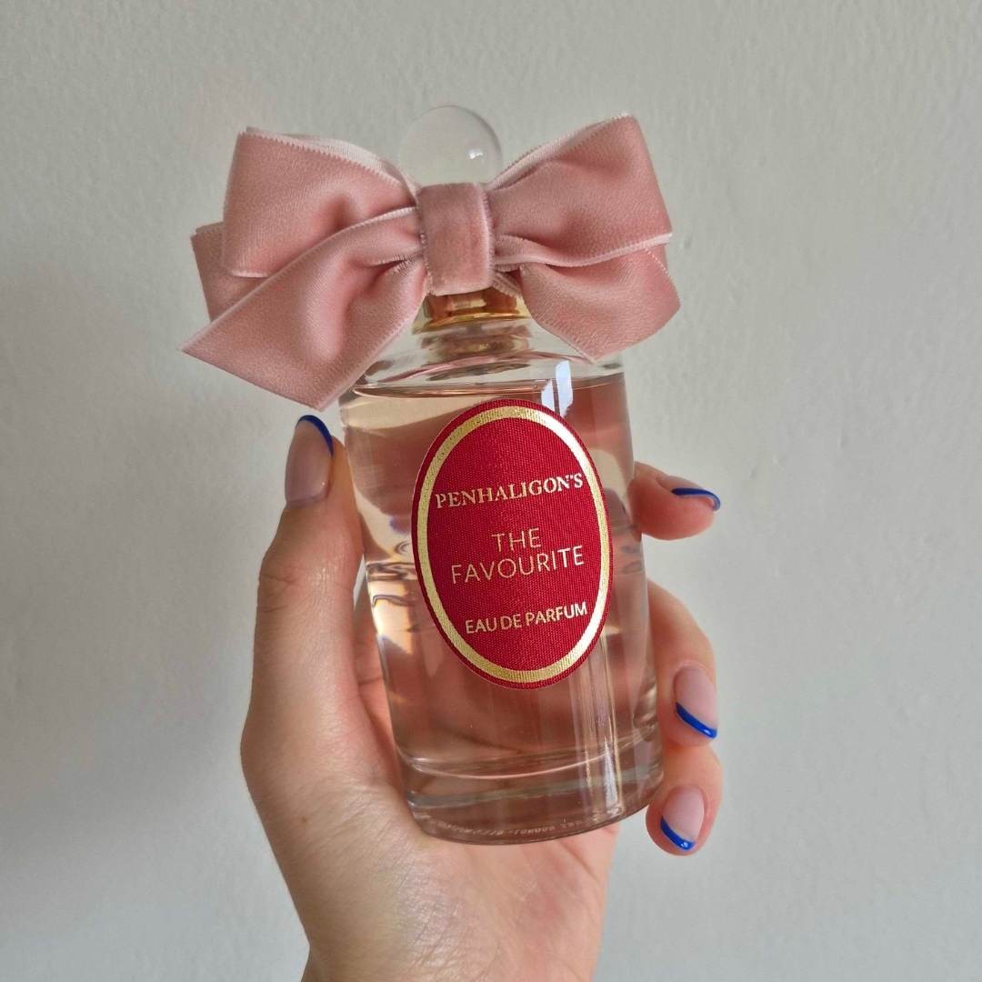 A close-up of Digital Beauty Writer Naomi Jamieson's hand, seen holding a bottle of Penhaligon's The Favourite perfume, which features a pink velvet bow/ in front of a cream wall background