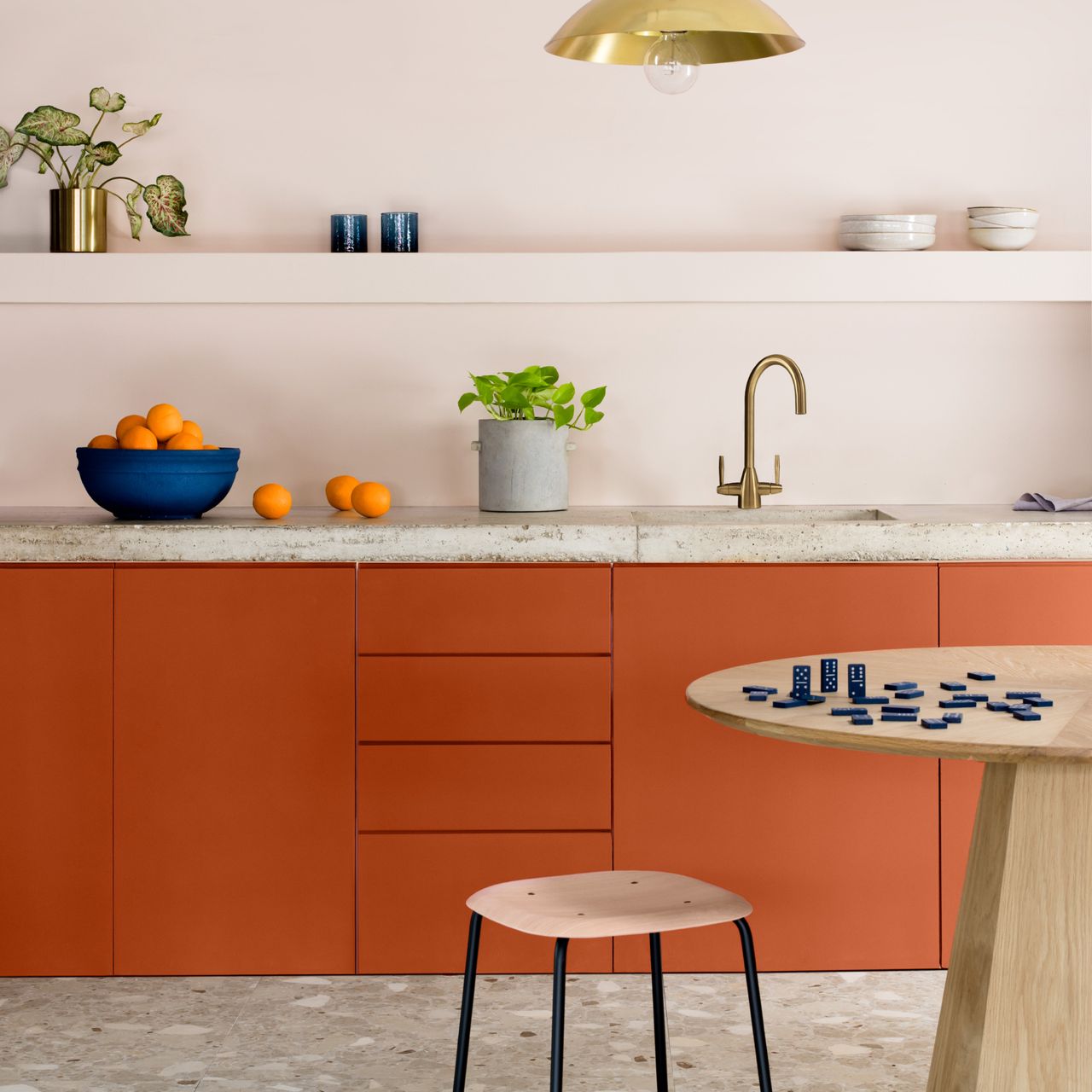 accent colour ideas, kitchen with pale blush walls, concrete worktop, brass tap, orange cabinets, terrazzo floor, table and stool, open shelving 