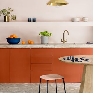 accent colour ideas, kitchen with pale blush walls, concrete worktop, brass tap, orange cabinets, terrazzo floor, table and stool, open shelving