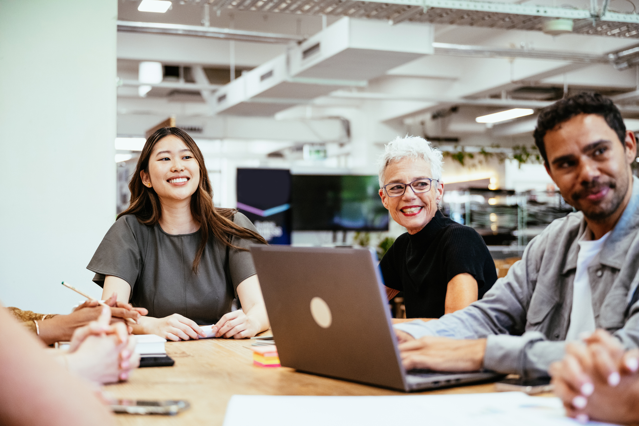 A diverse group of colleagues and business people collaborates on a project at a modern office table surrounded by plants and natural light.