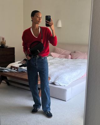 Woman wears a red Cashmere J. Crew sweater, black Adidas Samba sneakers, baggy jeans and a The Row belt.