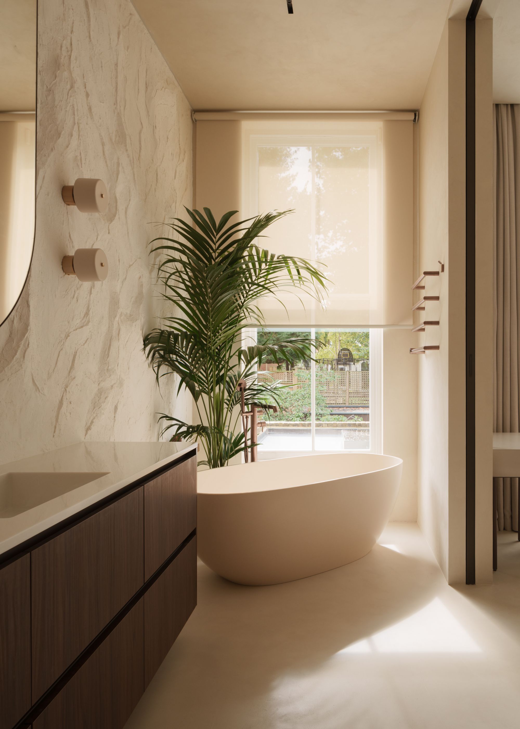 A modern bathroom with a textured cream wall, a dark wood vanity with a mirror above, and a bath and areca palm in the corner of the space in front of a window with a half-closed blind