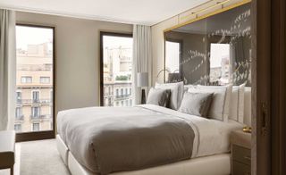 Interior view of a room at The One, Spain featuring light coloured walls, a bed with white and grey pillows and linen, a gold framed reflective headboard with a white feather style design, two tall windows offering a view of nearby buildings, white curtains, a bench style seat, a bedside table and a lamp