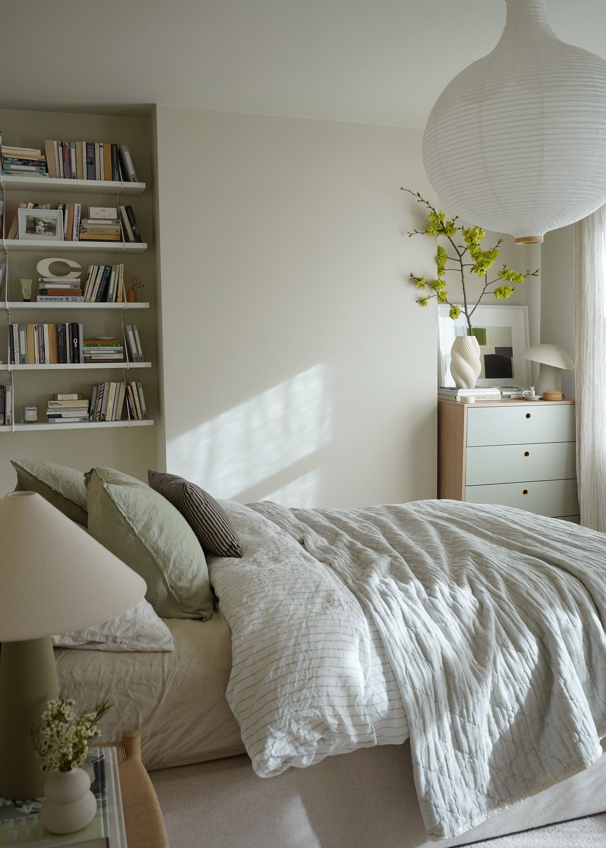 A neutral scandi-style bedroom with neutral layered bedding and green cushions, white paper ceiling light, and built-in bookshelves.