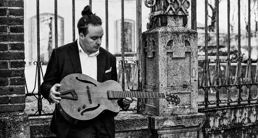 A black-and-white portrait of jazz guitarist Andrea Rinciari examining his instrument, as he stands in front of a metal gate.