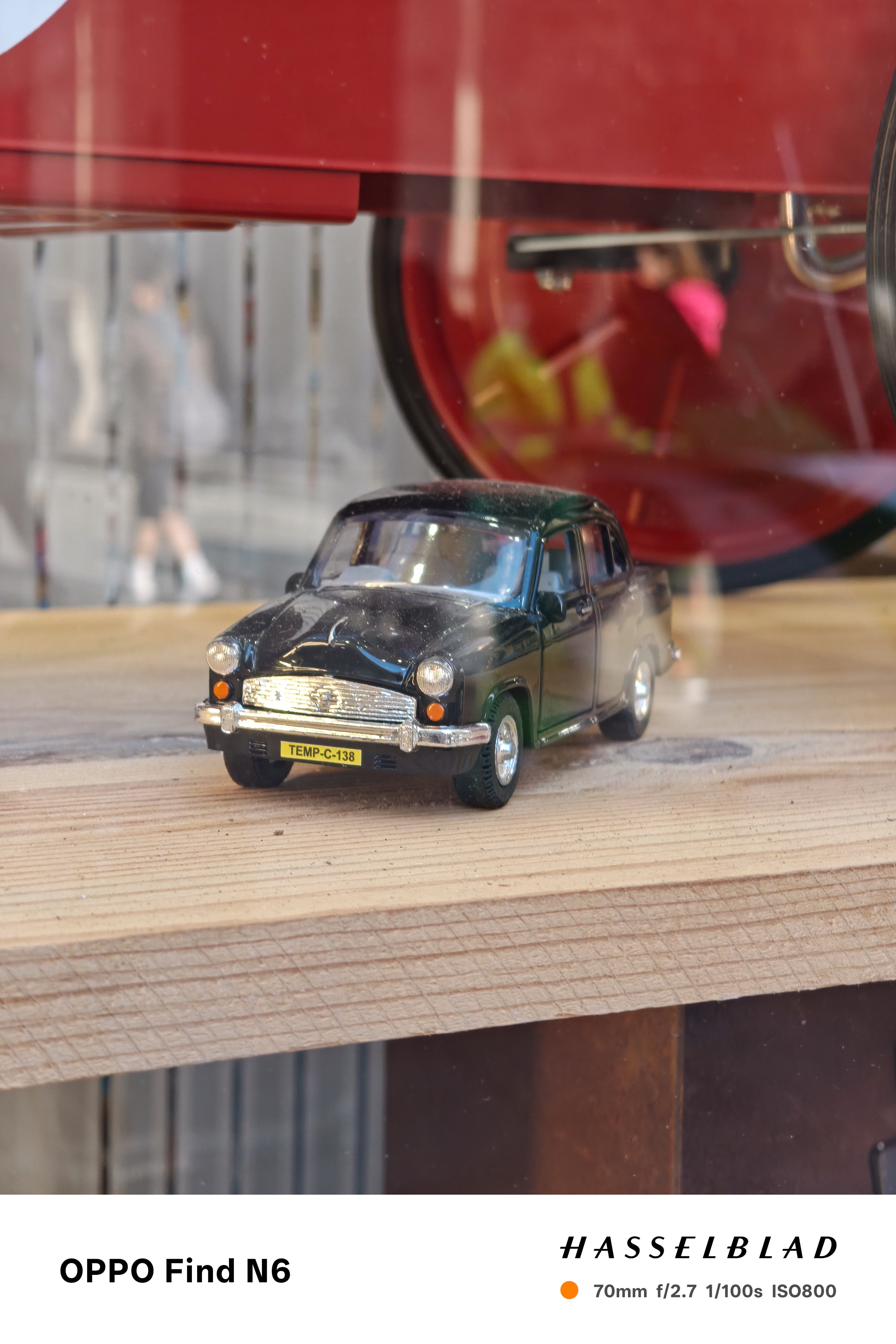 A close-up photograph of a vintage-style black toy car with silver chrome bumpers and a yellow license plate that reads "TEMP-C-138." The car is resting on a light wooden shelf, with a soft, blurred reflection visible in the glass behind it.