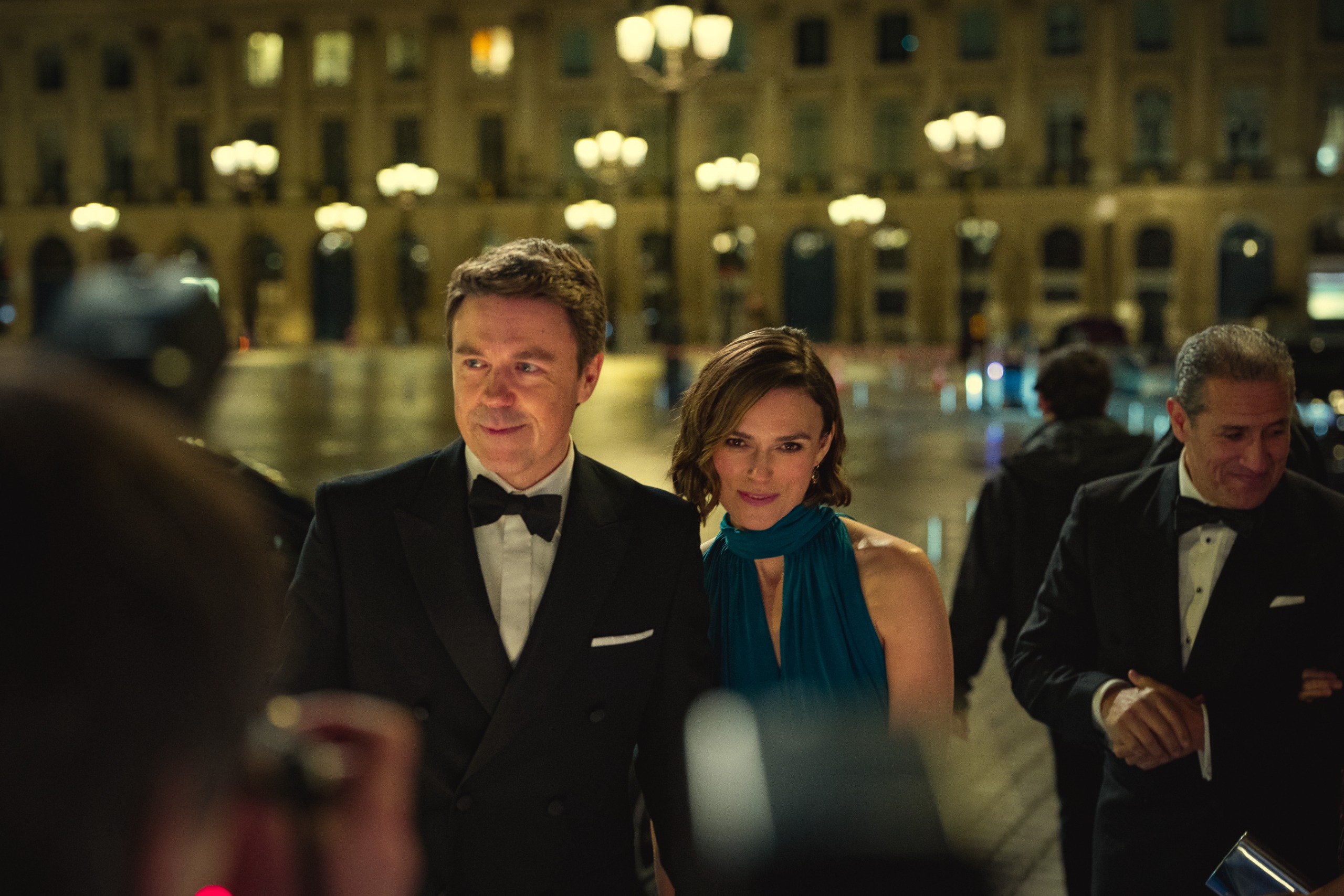 Andrew Buchan as wallace and keira knightley as helen wearing formal dress as they walk through a crowd of paparazzi in london at night in black doves season 2