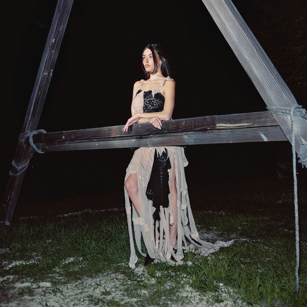 Image from Madison Beer's Who What Wear February cover story. It's dark out and she is leaning against a wood A-frame structure wearing a nude and black spaghetti strap dress with fringe detailing at the bottom. 