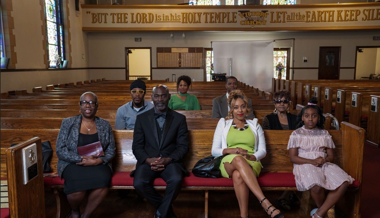African-American parishioners sit in pews in a Baptist church, facing the front.